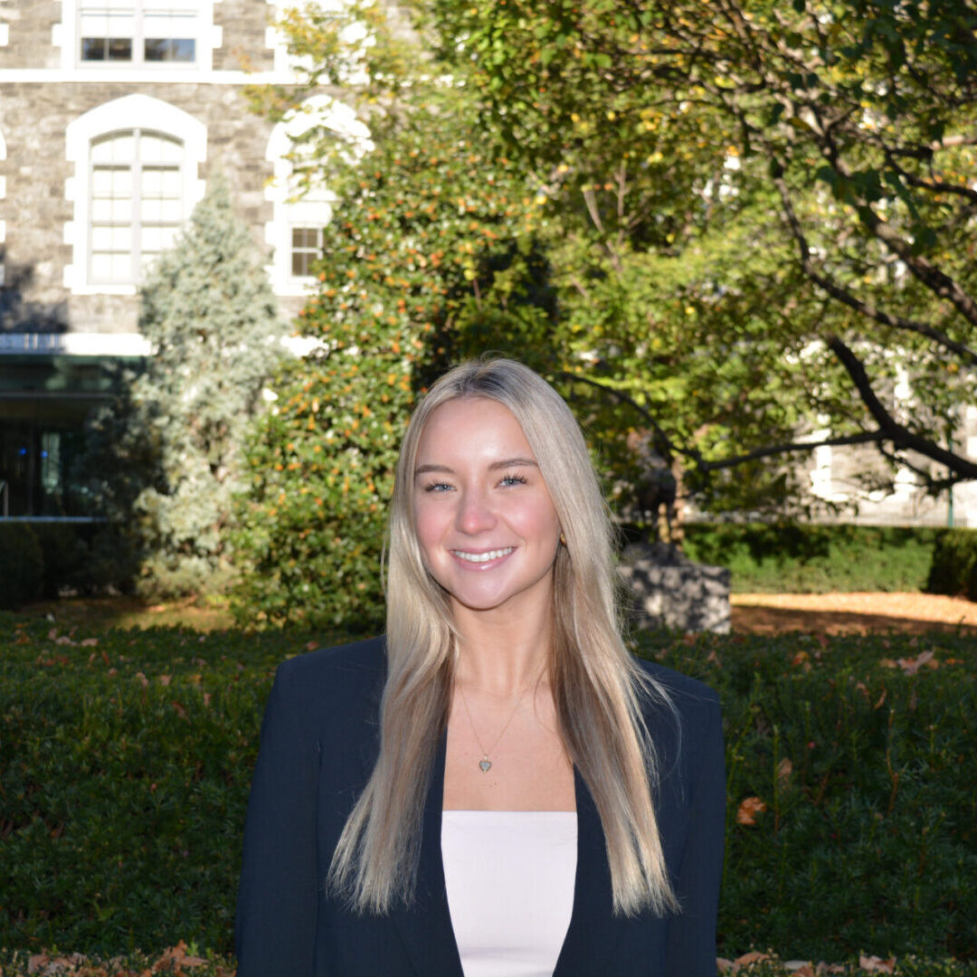 Smiling young woman outdoors with greenery and buildings in background.