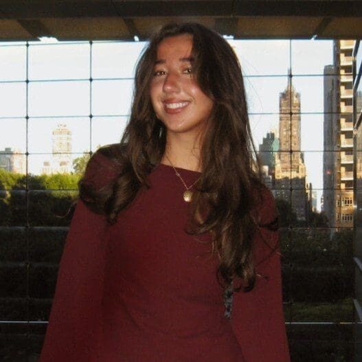 A young woman smiling indoors with city buildings in the background.