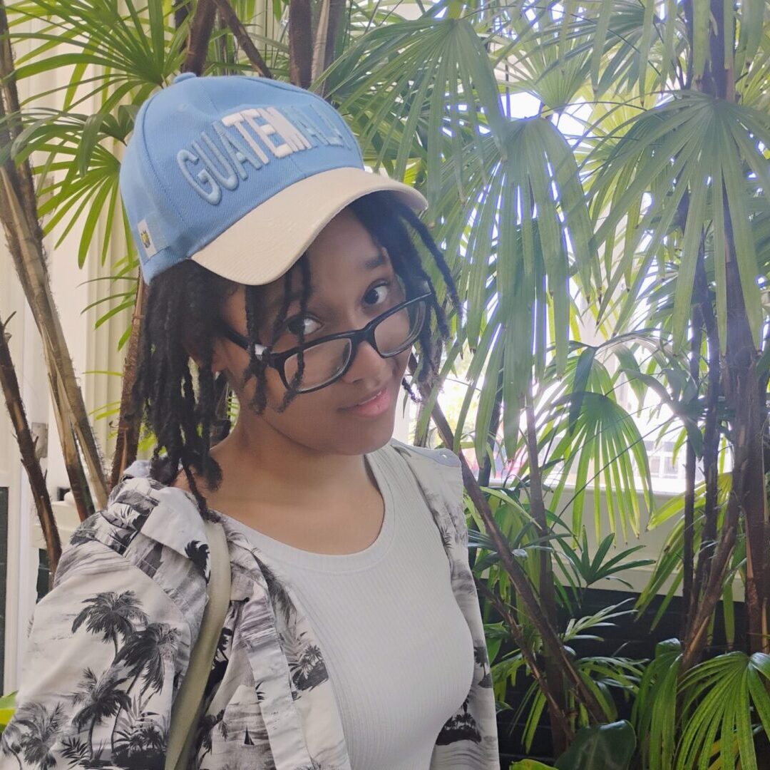 Person wearing glasses, a blue cap, and a patterned shirt, surrounded by lush green plants.