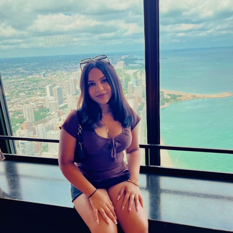 A woman sitting by a large window with a scenic city and ocean view.