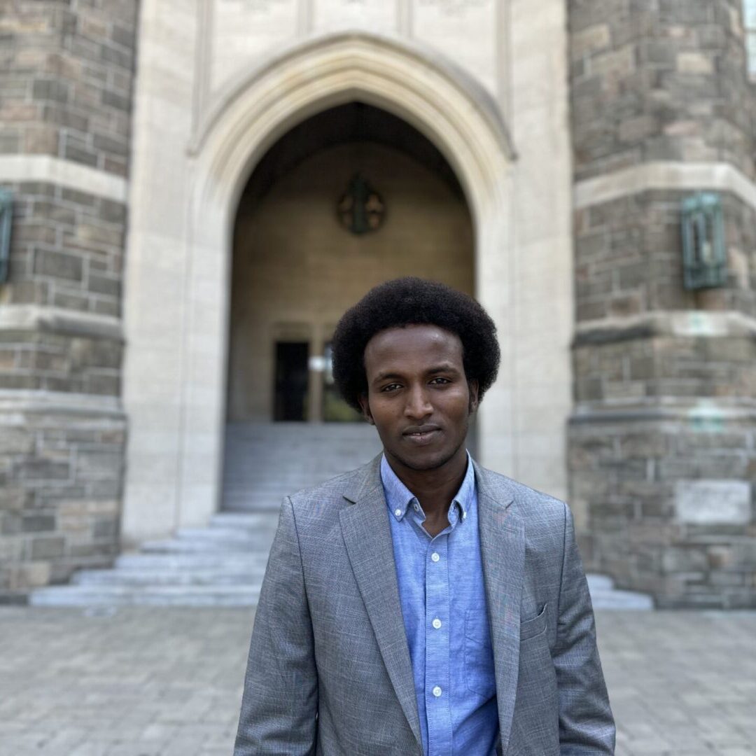 Man in a gray blazer stands outside a historic stone building.