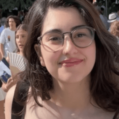Young woman with glasses smiling outdoors in a crowd.