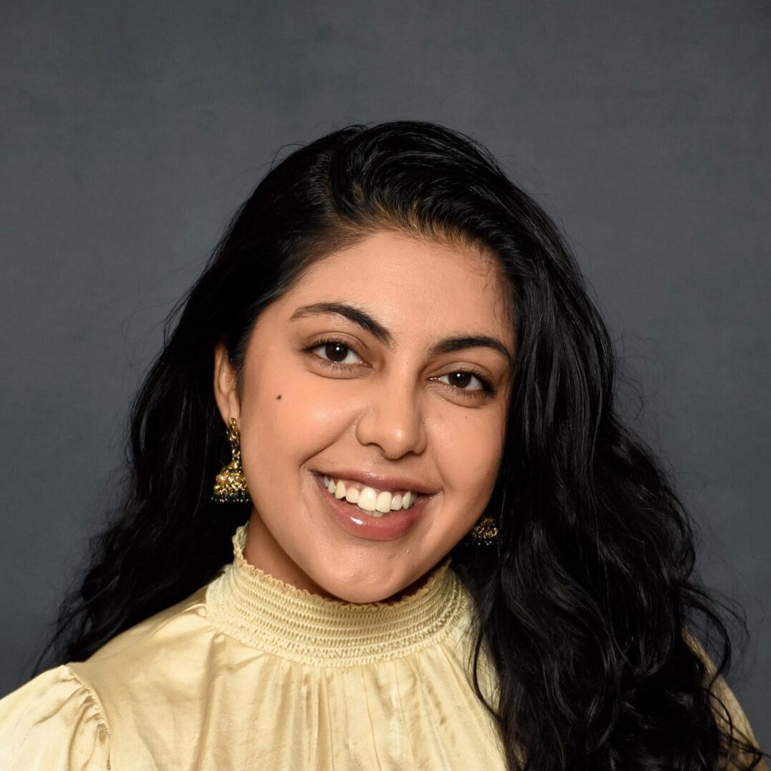 Smiling woman with long dark hair and cream blouse against a dark background.