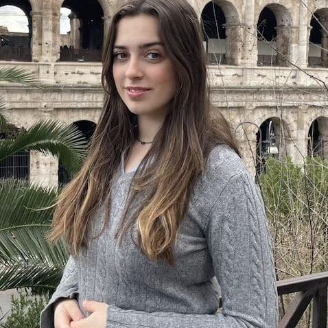 Young woman in a gray sweater poses outdoors with the Colosseum in the background.