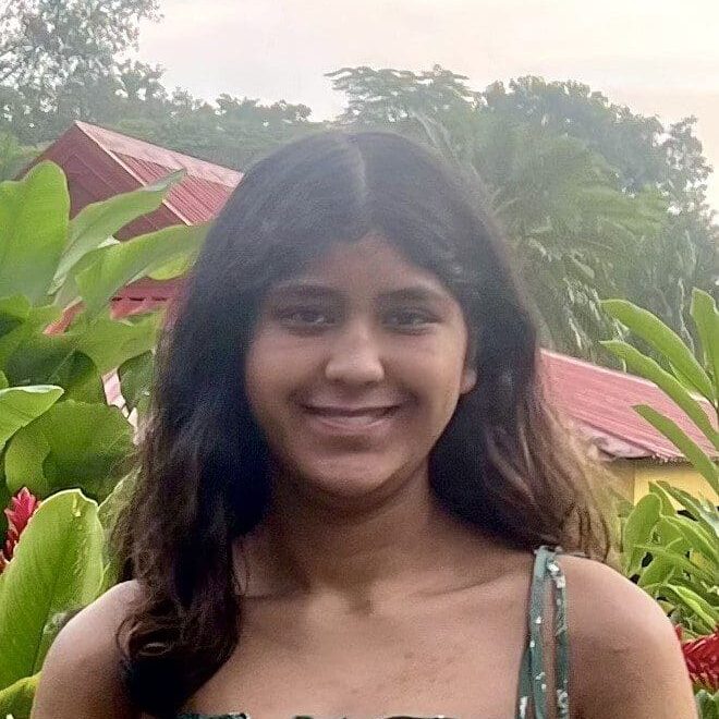 Smiling young woman outdoors with greenery and red-roofed buildings behind.