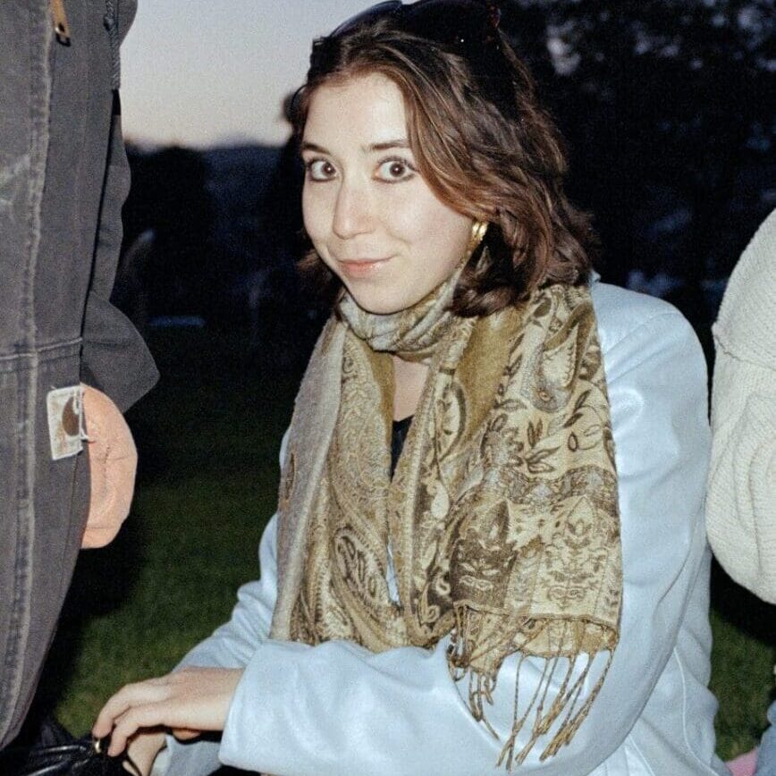 A woman with a golden scarf poses outdoors near a wooden structure at dusk.