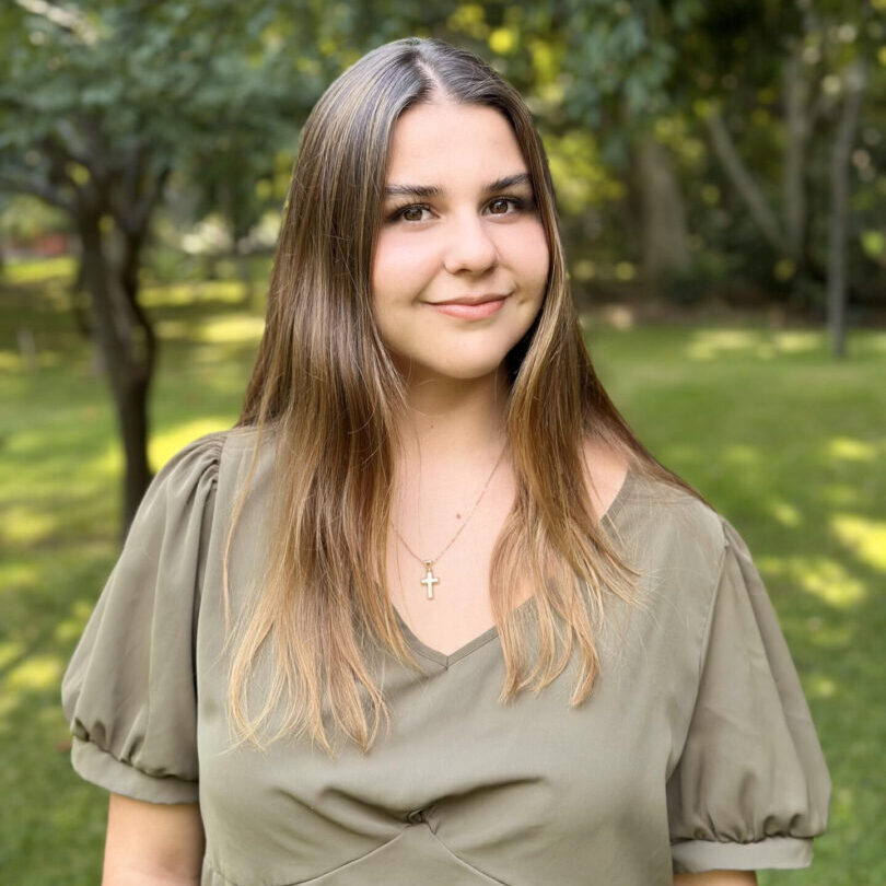 Young woman smiling outdoors with greenery in the background.